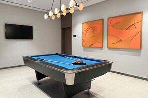 Une table de billard recouverte d’un drap bleu, dotée d’un socle noir et argenté, et accompagnée d’un jeu de queues de billard. Deux tableaux abstraits ornent le mur, et un téléviseur à écran plat est fixé au-dessus de la table.