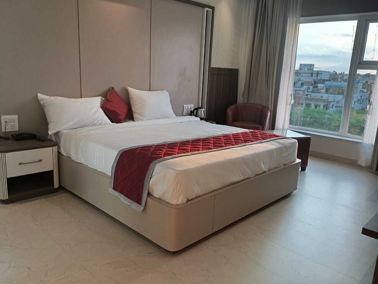 A modern hotel room with a large bed, a nightstand, a chair, and a view of the city through the window.