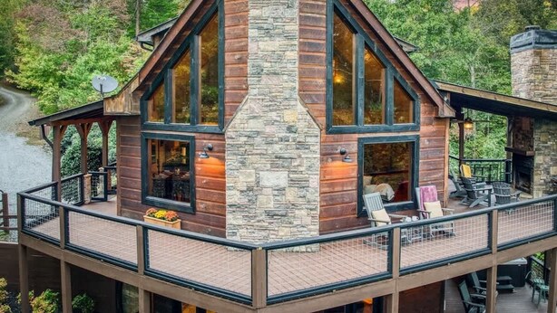 A cabin with a stone chimney, large windows, and a deck with outdoor seating.