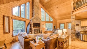 A log cabin interior with a stone fireplace, wooden furniture, and large windows.