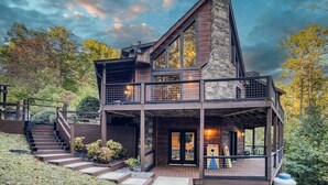 A two-story wooden cabin with a stone chimney, a balcony, and a staircase leading to the entrance.