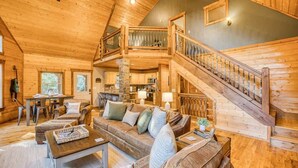 A spacious living room with a high ceiling, wooden floors, a staircase, and a kitchen in the background.