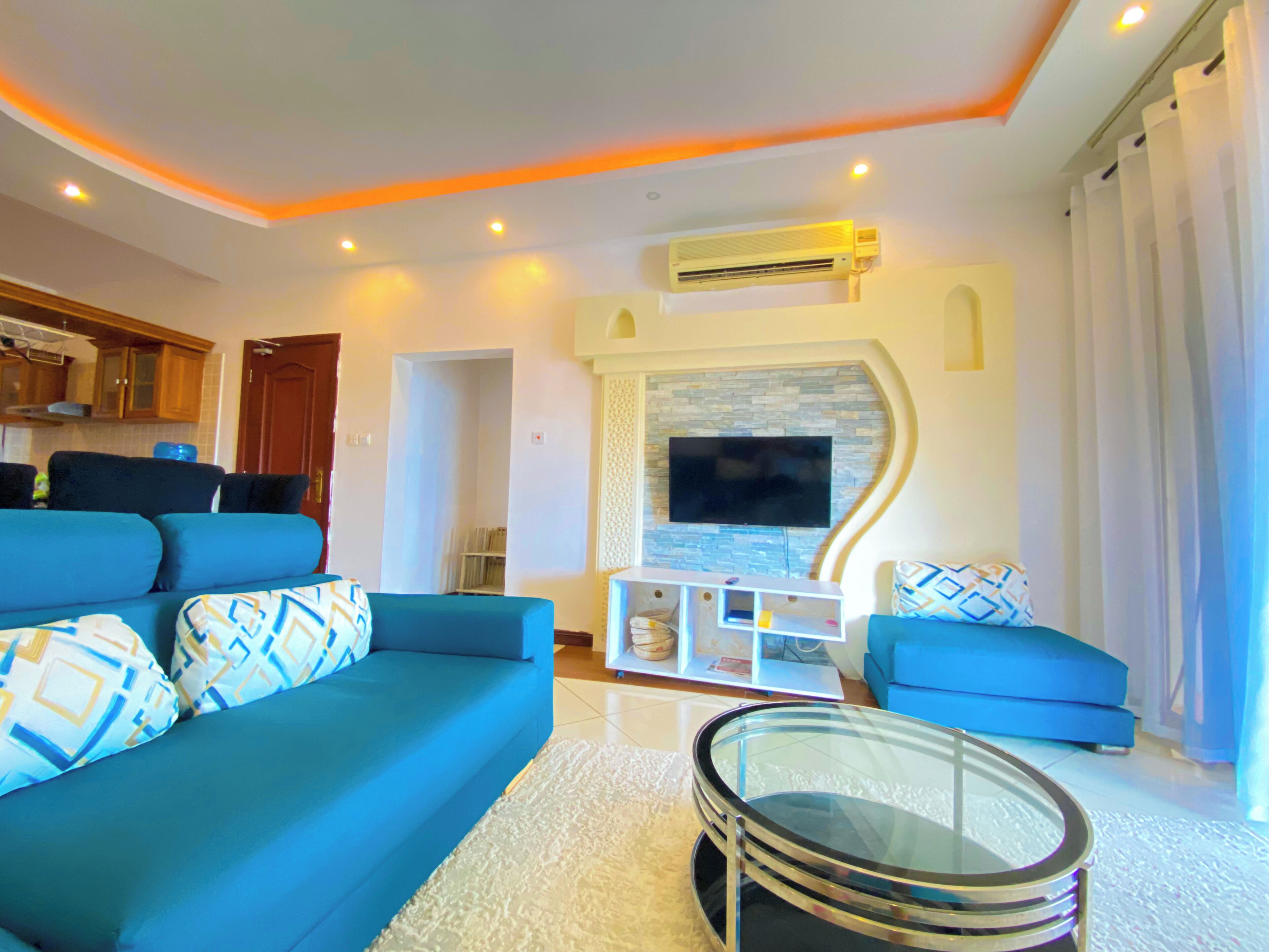 A modern living room with a blue sofa set, a glass coffee table, and a flat-screen TV mounted on a stone wall.