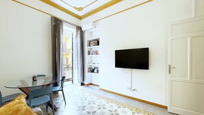 Un salon avec une table en verre, des chaises bleues, un canapé avec un coussin marron, un téléviseur à écran plat fixé au mur, une bibliothèque et un tapis à motifs.