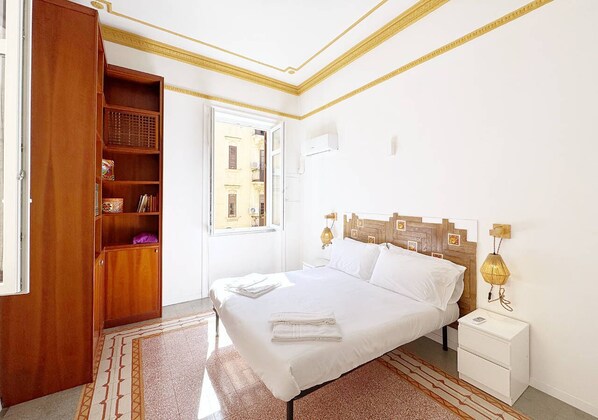 A bedroom with a large bed, a wooden wardrobe, a nightstand, and a window with a view of buildings.