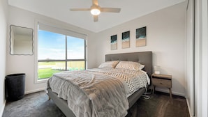 A bedroom with a large bed, a ceiling fan, a nightstand, and a window with a view of a grassy area.