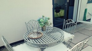 Una mesa redonda de metal con sillas blancas acolchadas, una cesta tejida y una planta en maceta en un patio con baldosas de piedra.