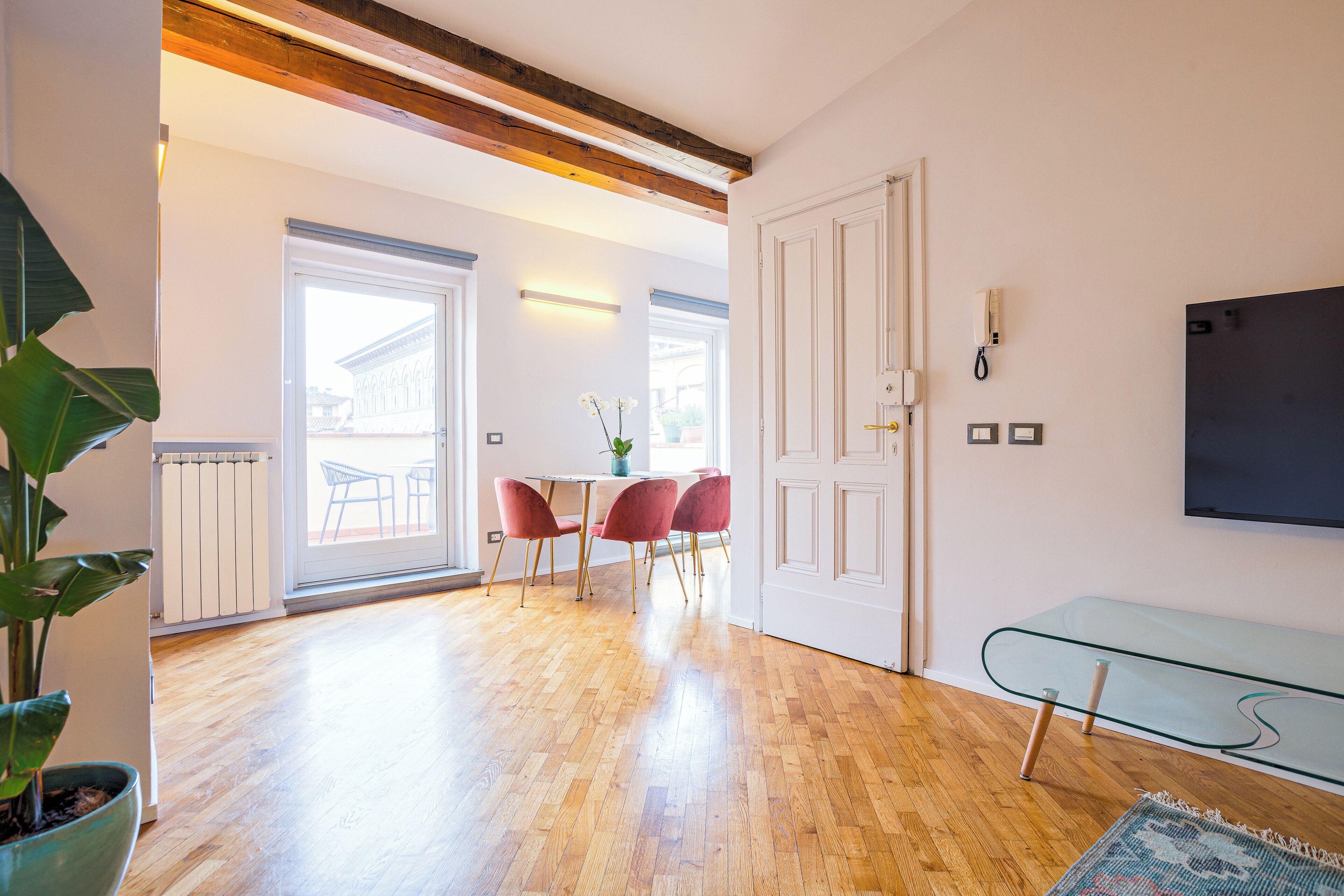 Une pièce avec un plancher en bois, un coin repas avec des chaises rouges, une porte blanche et une table en verre.