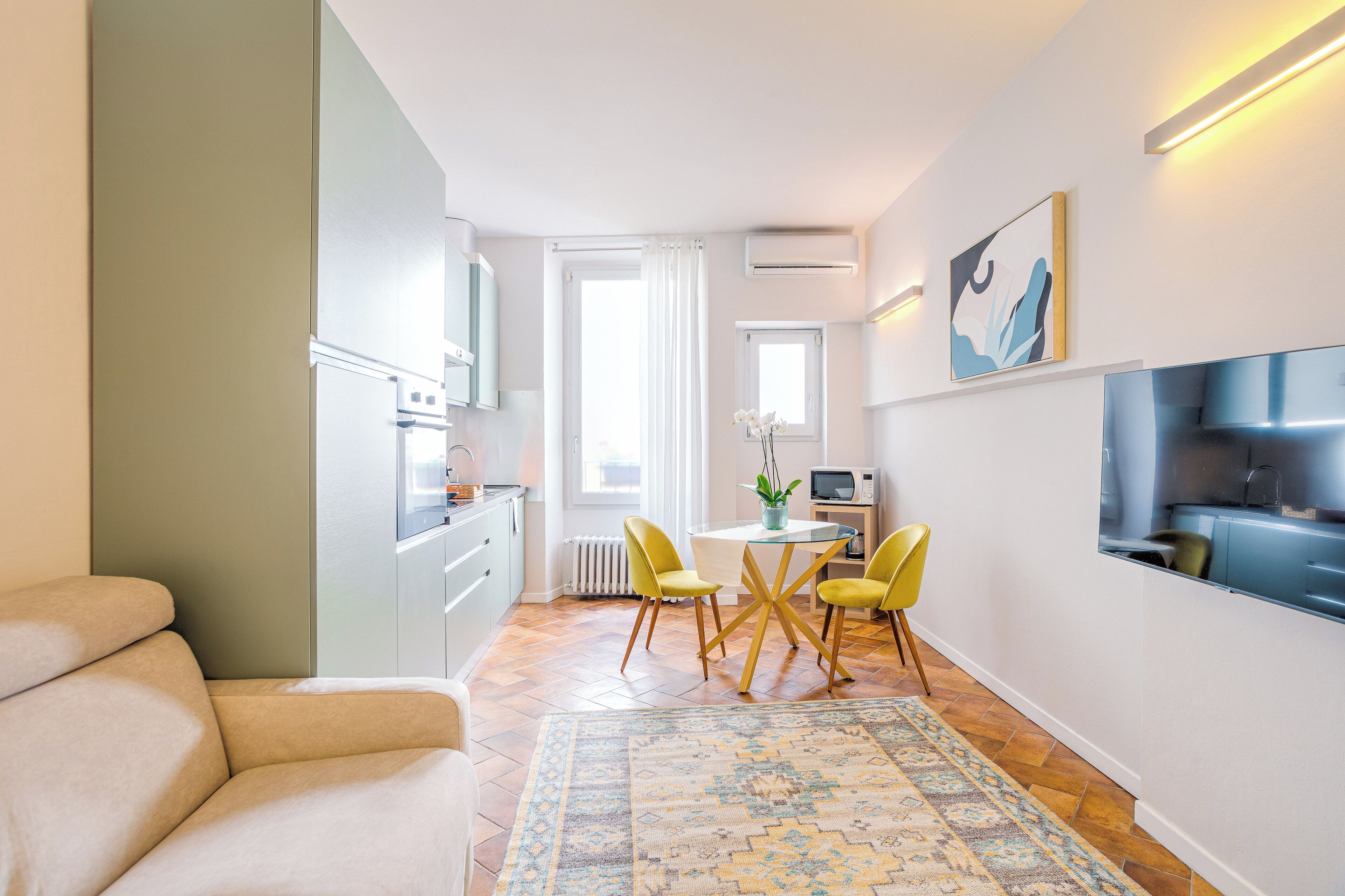 A modern living room with a sofa, a dining area with yellow chairs, a patterned rug, and a kitchen with white cabinets and a TV.