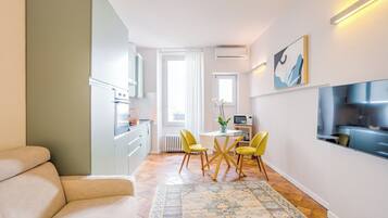 A modern living room with a sofa, a dining area with yellow chairs, a patterned rug, and a kitchen with white cabinets and a TV.