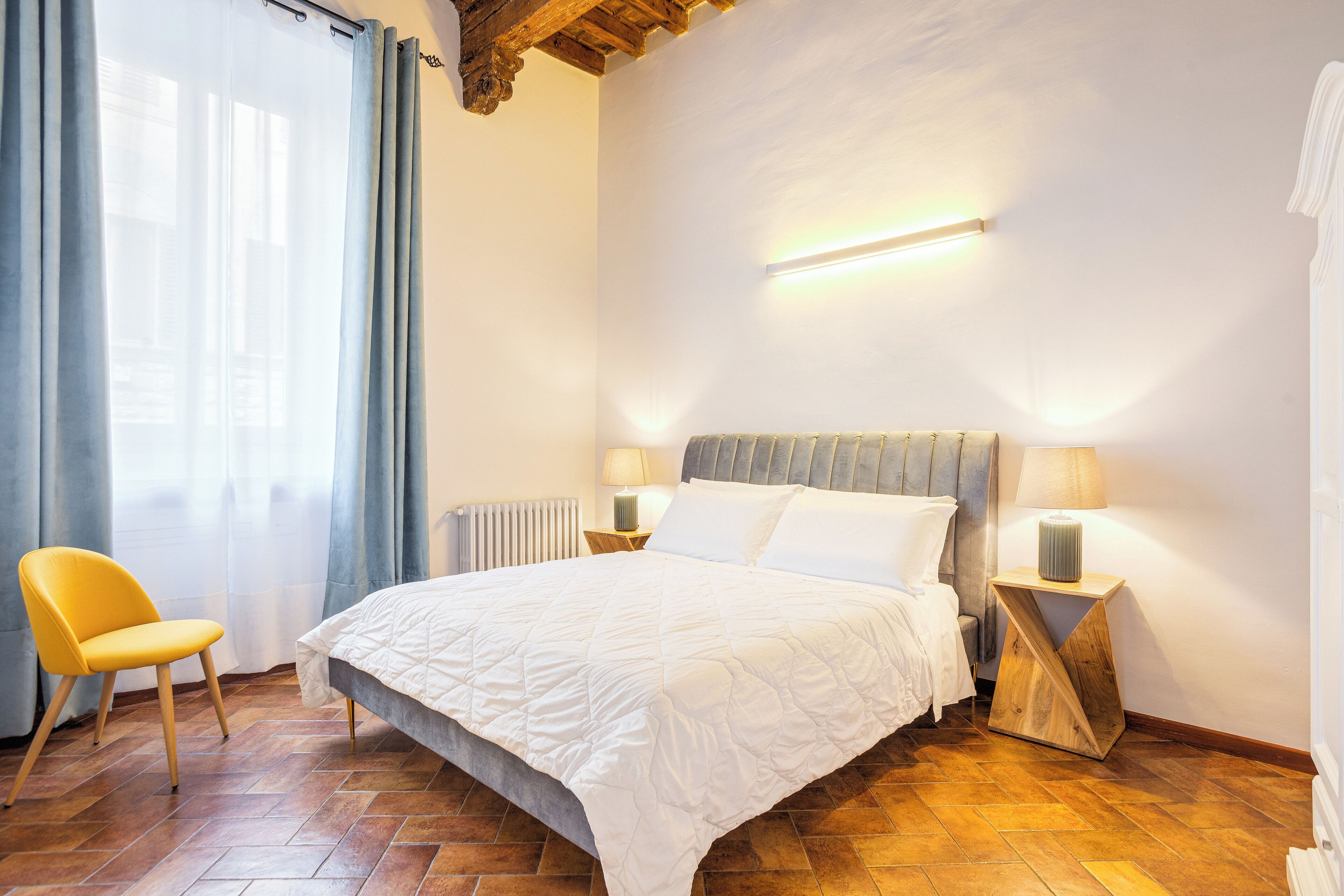 Un dormitorio con cama, una silla, una mesita de noche y una lámpara en la pared.