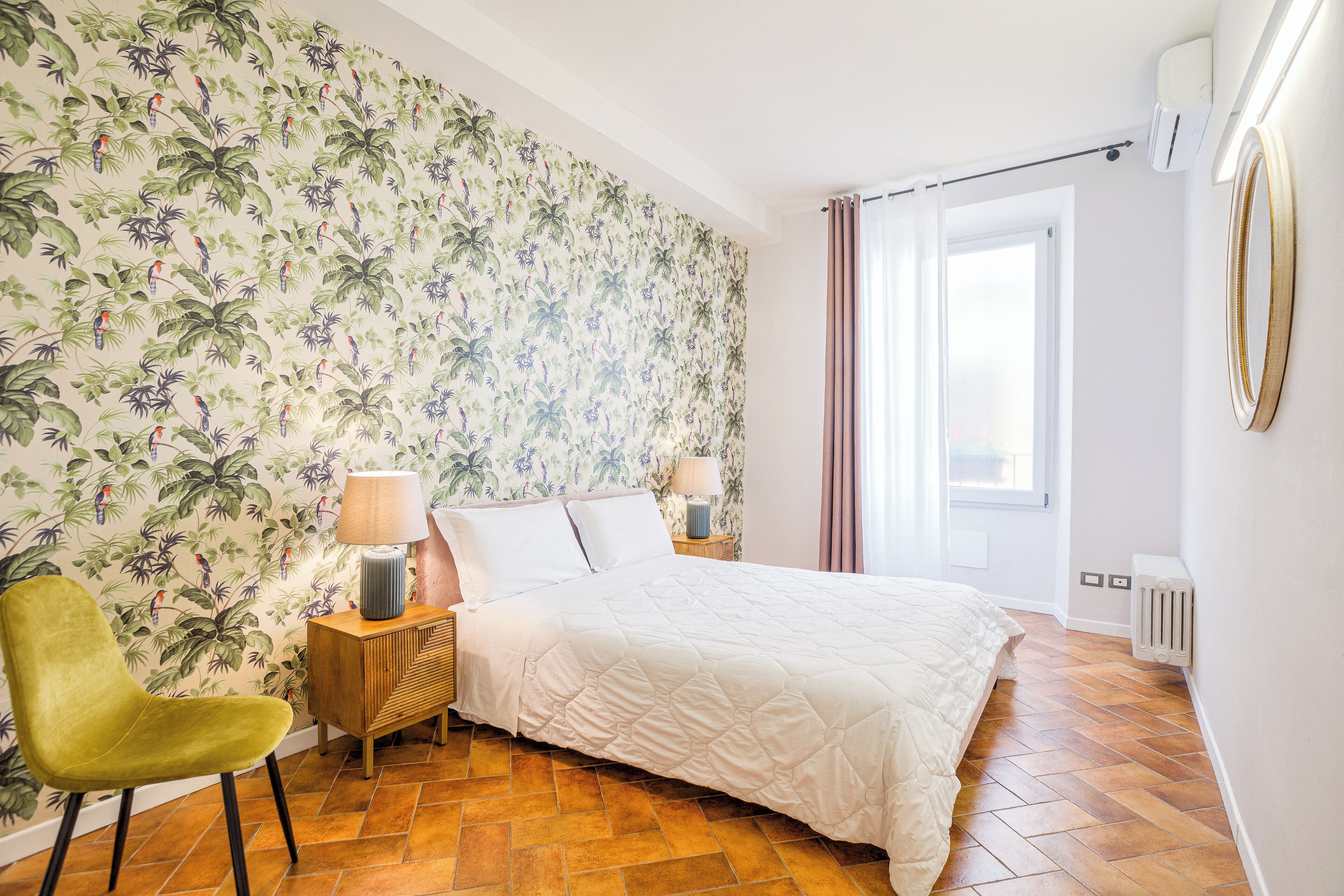 Une chambre avec un lit, un fauteuil vert, une table de chevet avec une lampe, un mur recouvert d’un papier peint à motifs tropicaux et une fenêtre avec des rideaux.