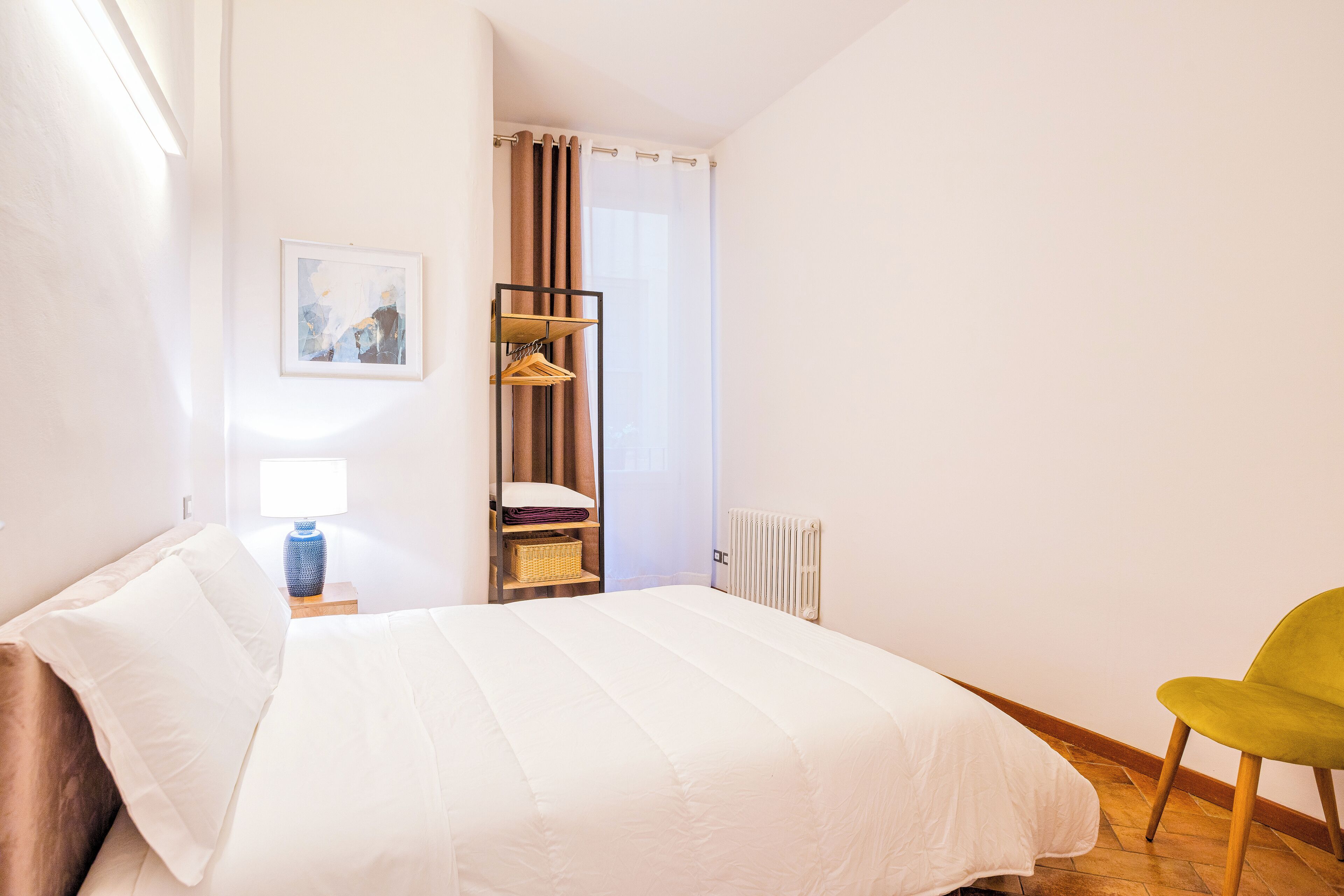 Un dormitorio con cama, mesita de noche, lámpara, silla y ventana con cortinas.