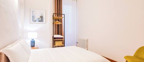 Une chambre à coucher avec un lit, une table de chevet, une lampe, une chaise et une fenêtre avec des rideaux.
