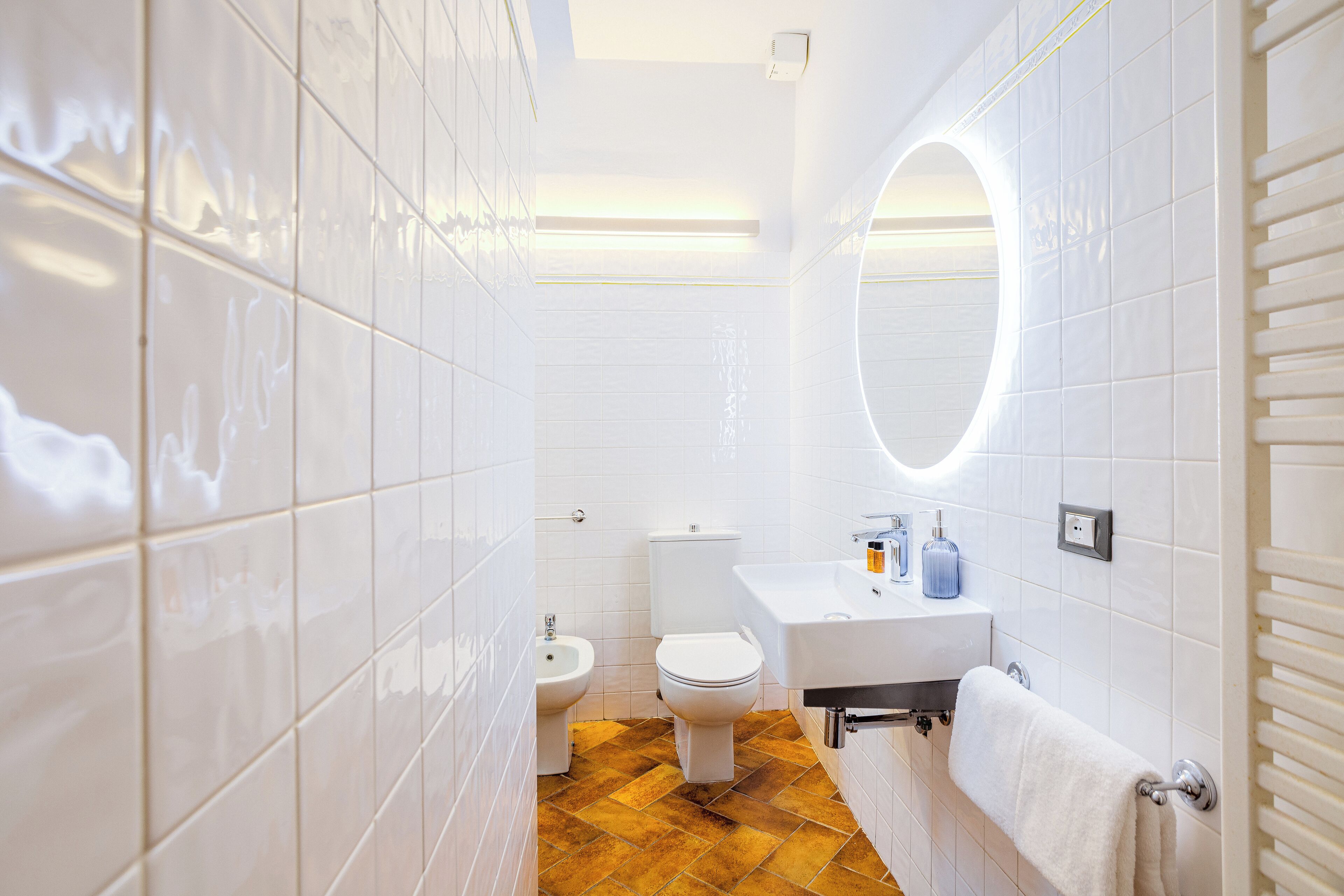 Un baño con azulejos blancos, un inodoro, un bidé, un lavabo con espejo y un perchero para toallas.