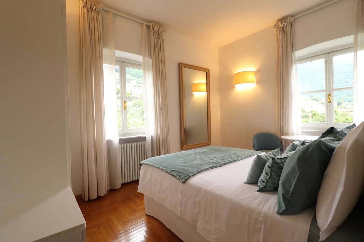 A bedroom with a bed, a chair, a table, a mirror, and a window with curtains.