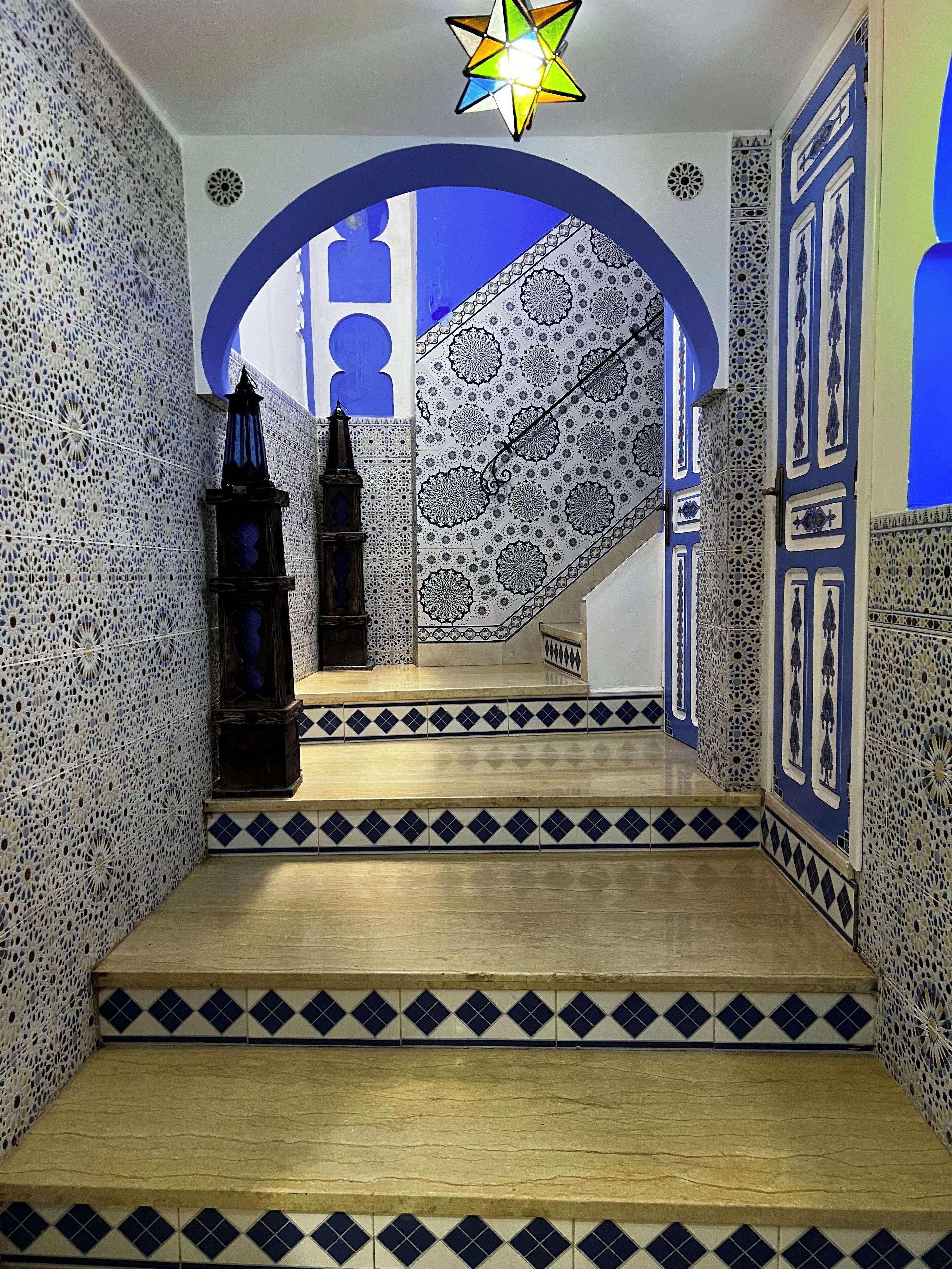 Un escalier recouvert de carreaux bleus et blancs formant un motif, menant à une pièce dont le mur est également décoré d’un motif bleu et blanc, et qui est éclairée par un luminaire en forme d’étoile.
