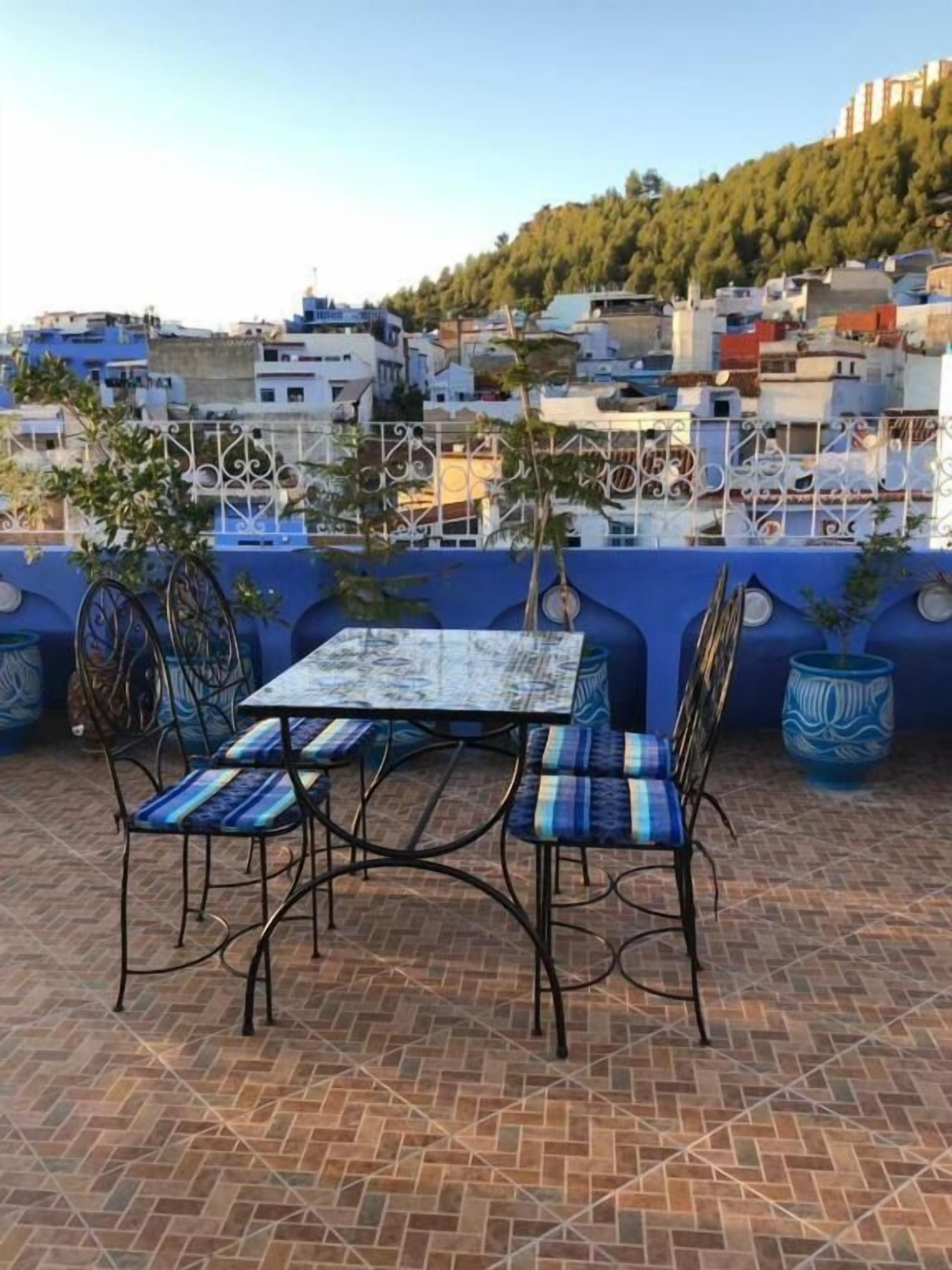 Eine Terrasse mit Tisch und Stühlen, umgeben von Blumentöpfen und Blick auf einen Hügel mit Gebäuden.