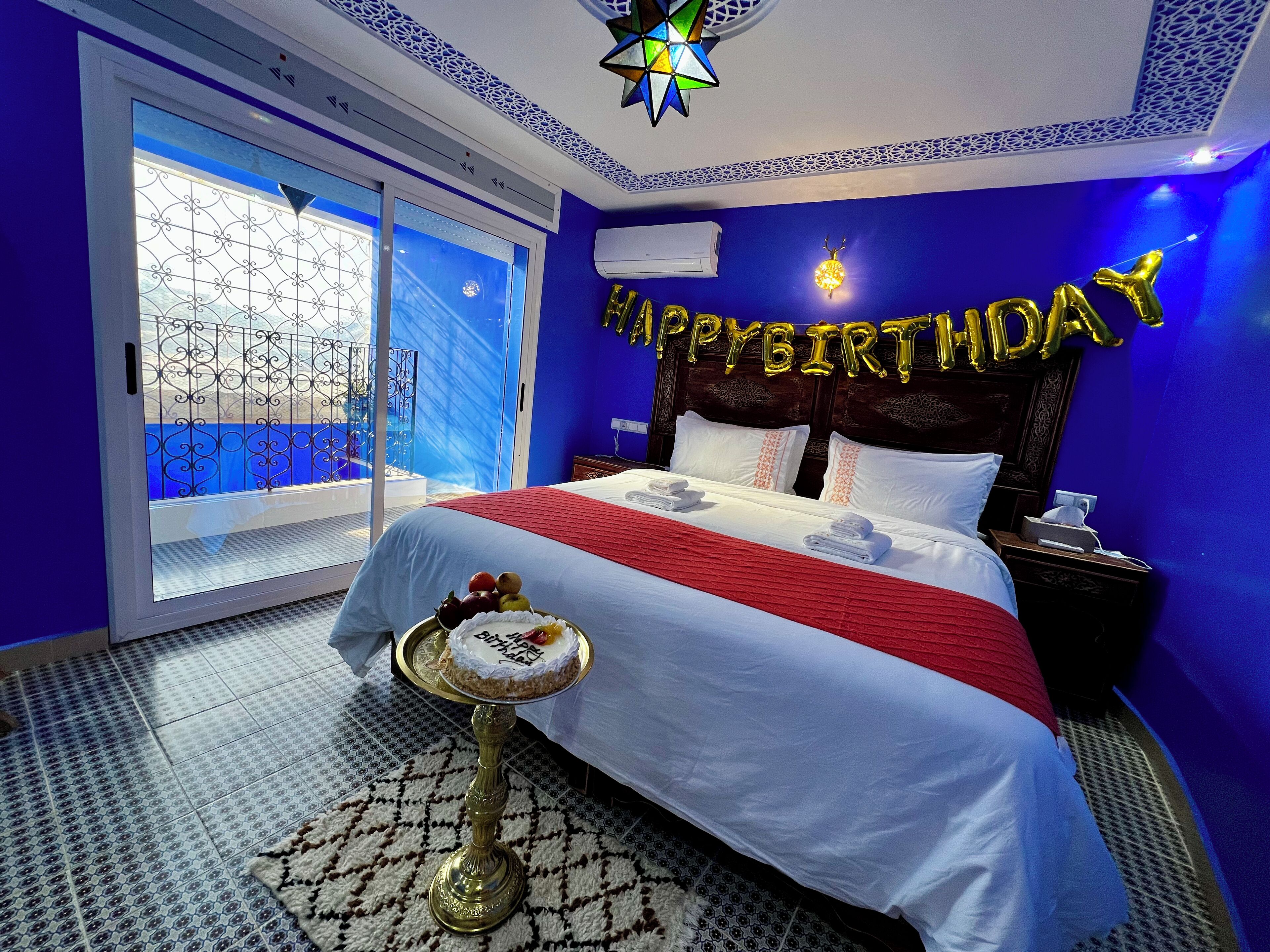 A hotel room with a bed, a bedside table with a cake, and a 'Happy Birthday' balloon decoration.