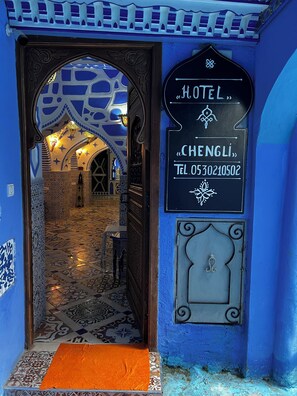 An ornate hotel entrance with a blue facade, a signboard displaying the name "HOTEL CHENGLI" and a contact number, and a patterned tiled floor leading into the interior.