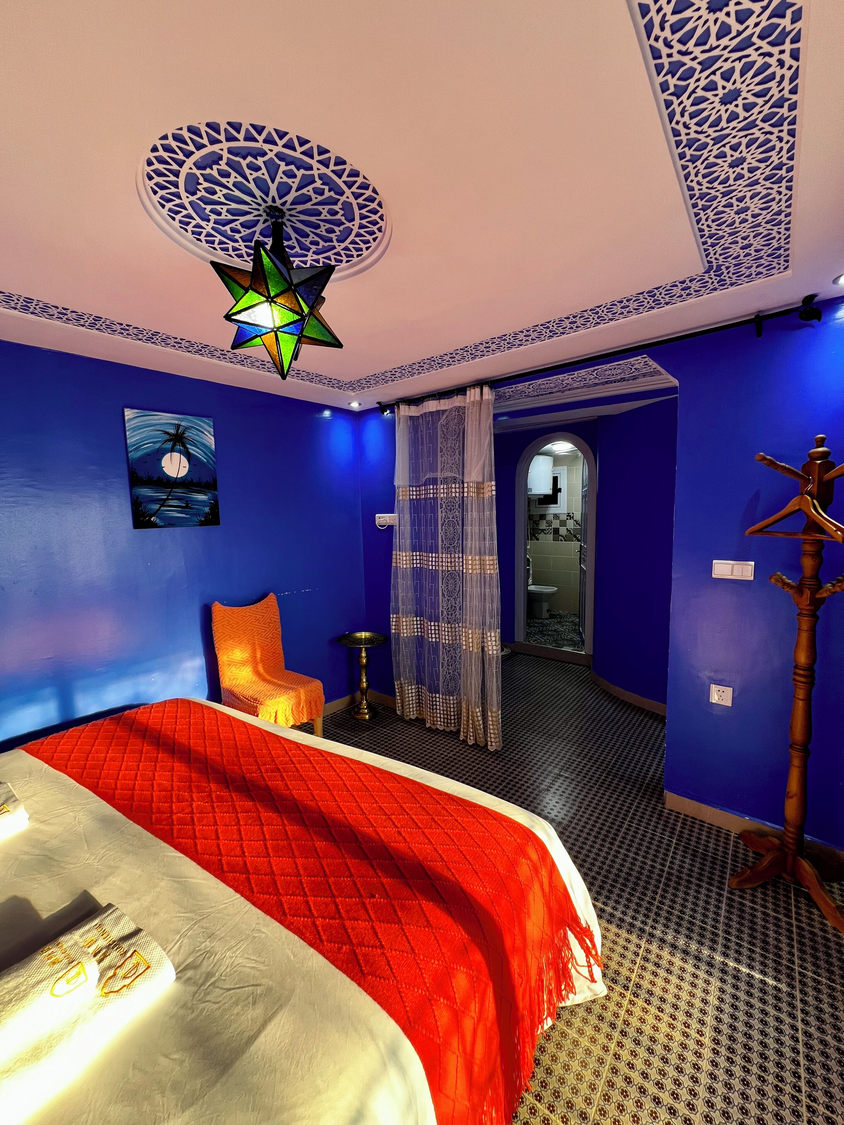 A room with a bed, a nightstand, a decorative ceiling light, and a view into another room.