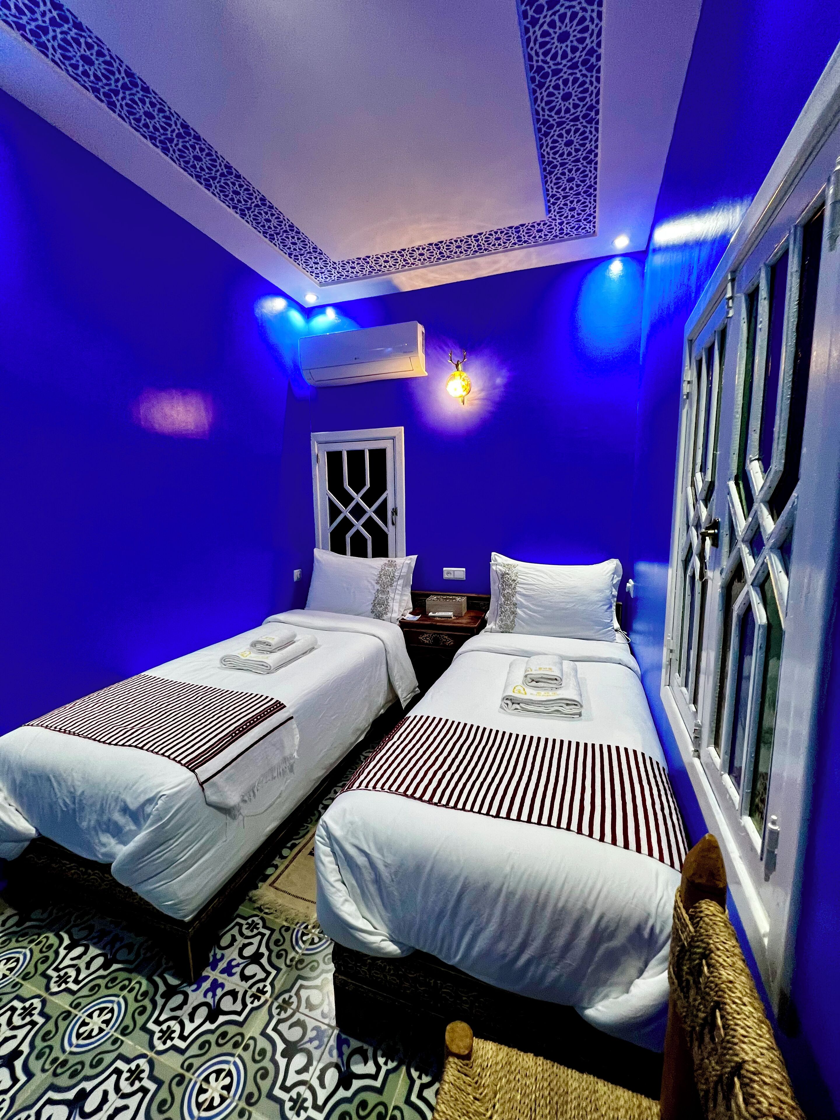 A room with two beds, a window with decorative iron bars, a wall-mounted air conditioner, and a patterned floor.