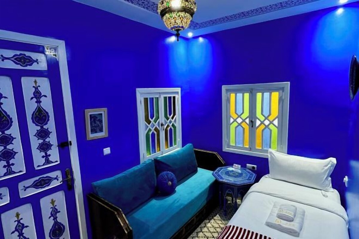 A room with a blue wall, a sofa, a bed, and a chandelier.