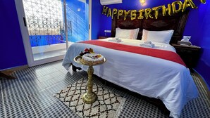 Habitació d'hotel amb un llit, una taula de nit amb un pastís, una decoració de globus que diu 'HAPPY BIRTHDAY' i una vista de l'exterior a través d'una porta de vidre.