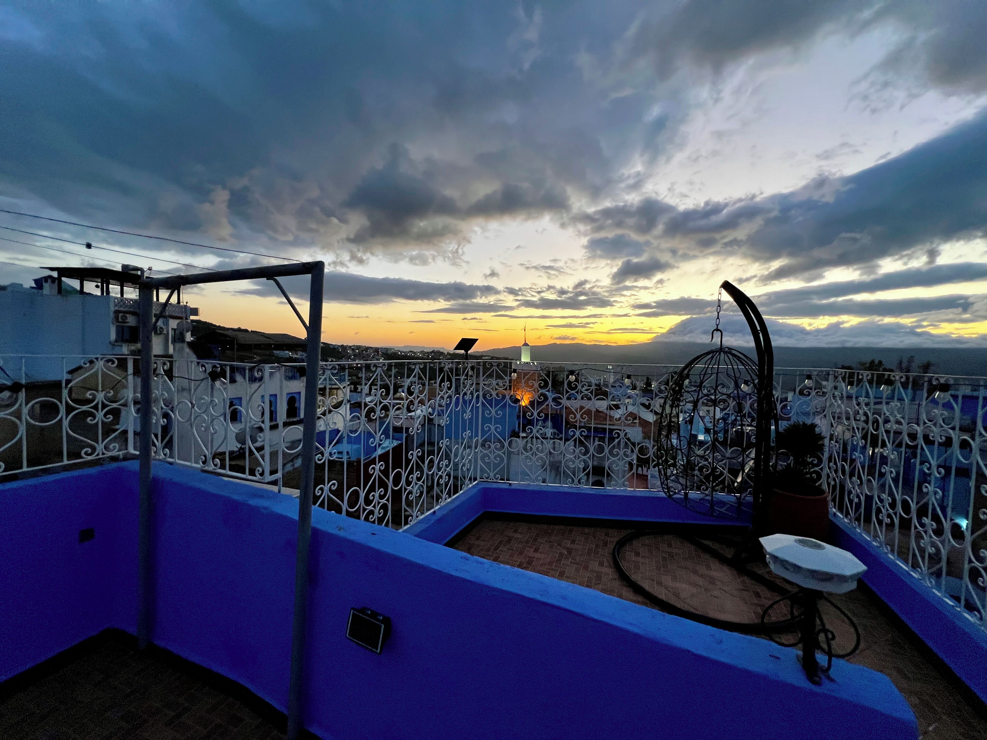 Une terrasse sur le toit offrant une vue sur les toits de la ville au coucher du soleil, avec une rambarde bleue et une clôture en métal décorative.
