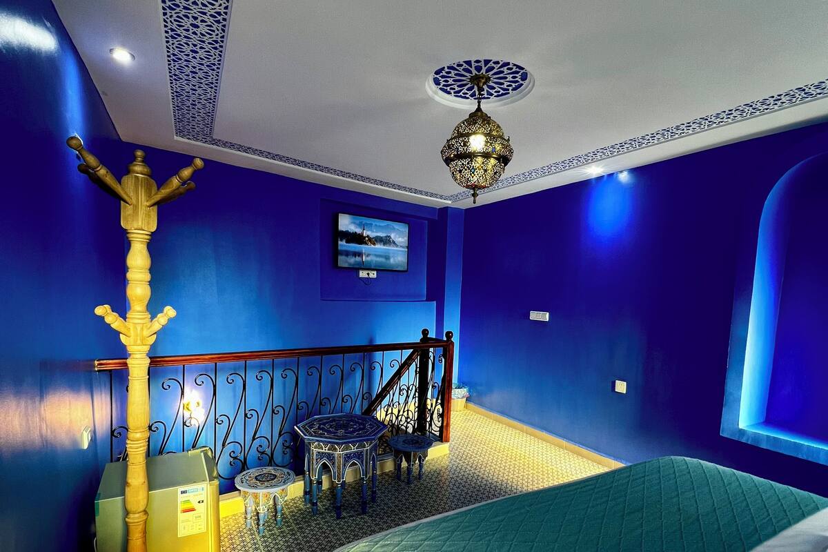 Ein Zimmer mit einem Bett, einem Sessel, einem kleinen Tisch, einer dekorativen Stehlampe und einem an der Wand befestigten Fernseher.