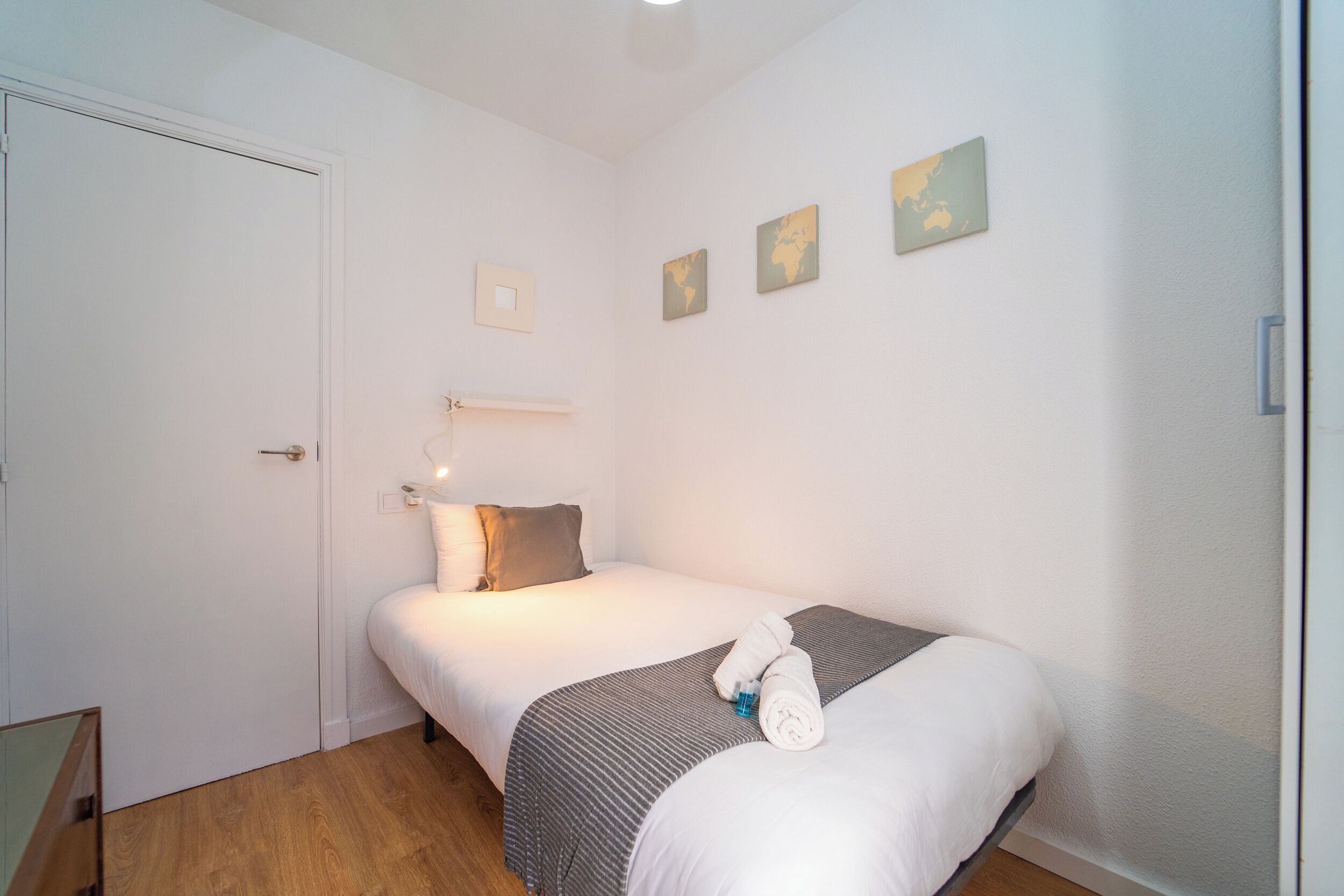 Ein Einzelbett mit grauen und weißen Streifen, ein weißes Kopfkissen und ein zusammengerolltes Handtuch darauf. Darüber an der Wand hängen drei gerahmte Bilder.