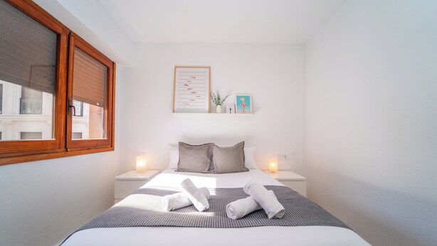 Cama bem arrumada com lençóis brancos e manta cinza listrada, duas toalhas brancas, uma lista emoldurada na parede e uma pequena planta em vaso.