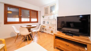Uma sala de estar moderna com uma mesa de jantar de madeira, cadeiras brancas, uma TV de tela plana sobre um suporte de madeira e uma estante branca com objetos decorativos.