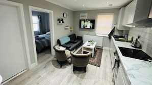 A modern living room with a sofa, two armchairs, a coffee table, and a TV. There is a kitchen area with a stove and cabinets. - Stylish and Luxury Flat in LND (Uxbridge)