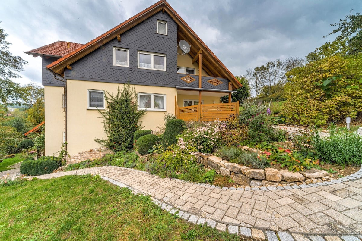 Ein zweistöckiges Haus mit Balkon, umgeben von einem Garten mit verschiedenen Pflanzen und einem Steinweg.