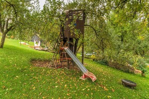 A playground with a slide, a swing set, and a small wooden structure in a grassy area with trees. - Cozy Apartment in Marktrodach With Sauna (Marktrodach)