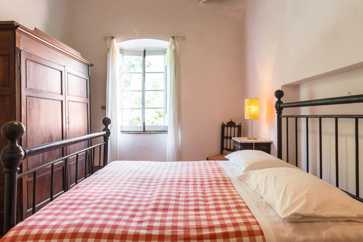 A bedroom with a checkered bedspread, a wooden wardrobe, a window with curtains, and a bedside lamp.