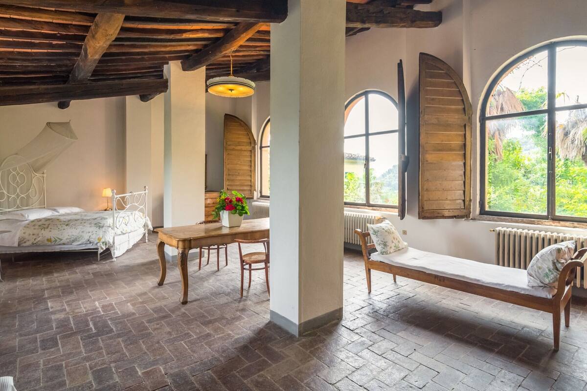 A room with a wooden ceiling, a bed with a canopy, a table with chairs, a bench, and large arched windows with a view of greenery.