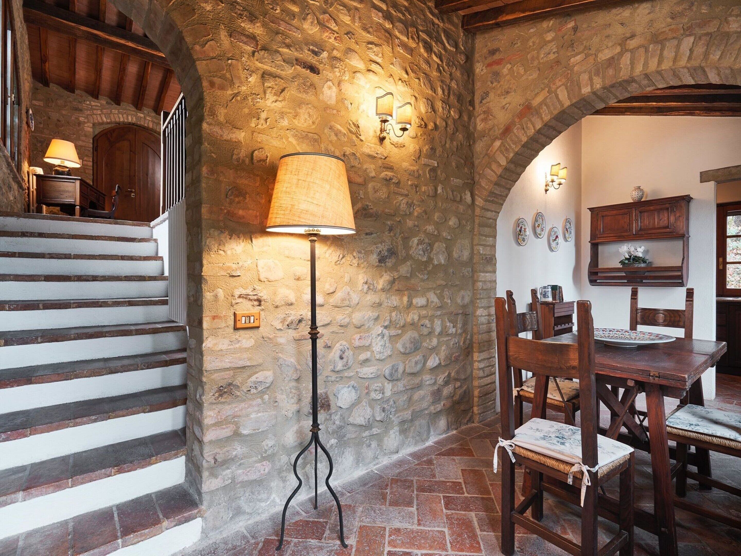 A rustic interior with stone walls, a wooden staircase, a dining table with chairs, and a floor lamp.