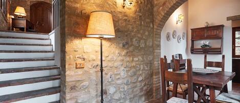 A rustic interior with stone walls, a wooden staircase, a dining table with chairs, and a floor lamp.