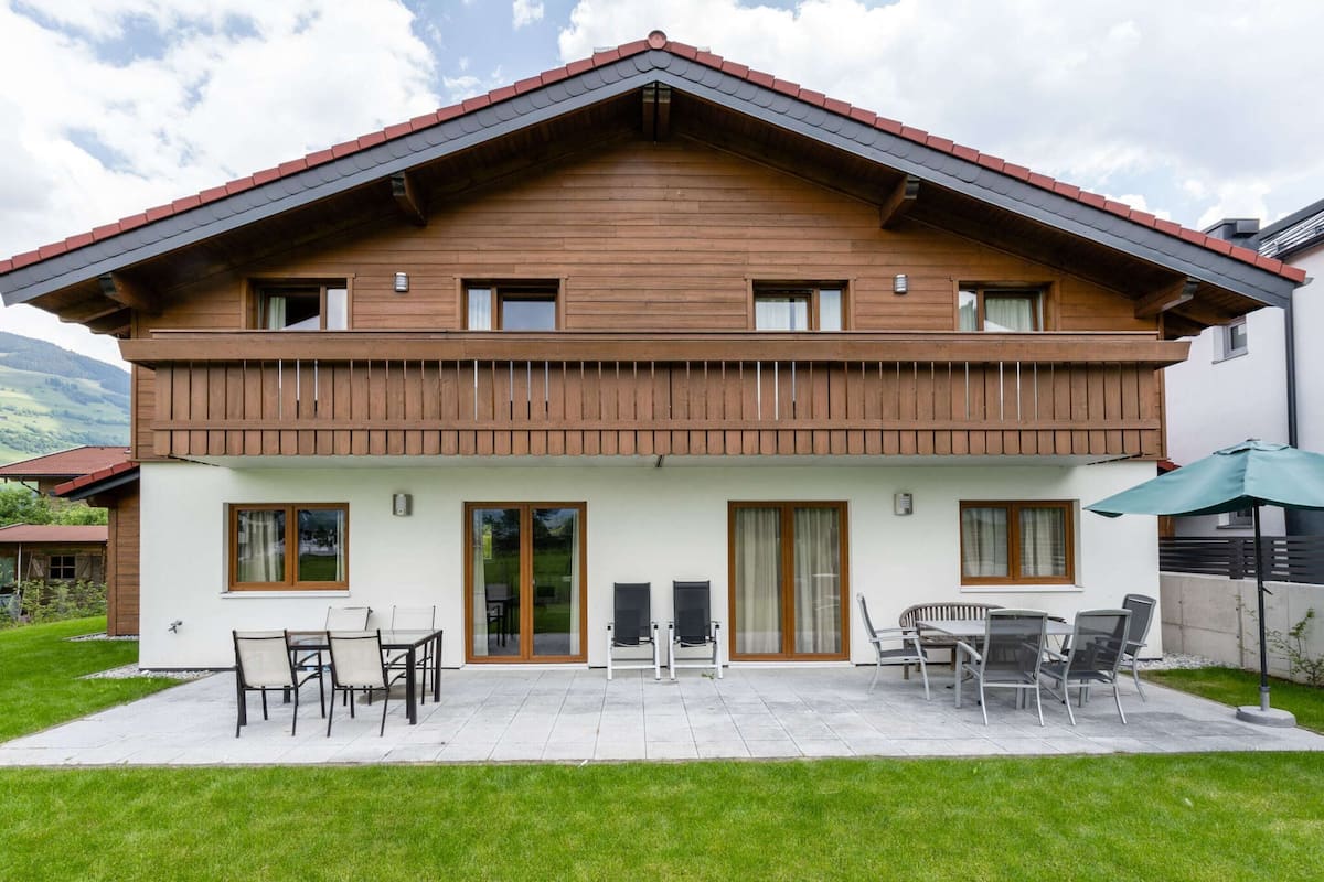 A two-story wooden house with a balcony, a patio with outdoor furniture, and a view of the surrounding landscape.