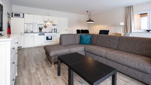 Un salón moderno con un sofá seccional gris, una mesa de centro negra y una zona de cocina con armarios blancos y un microondas.