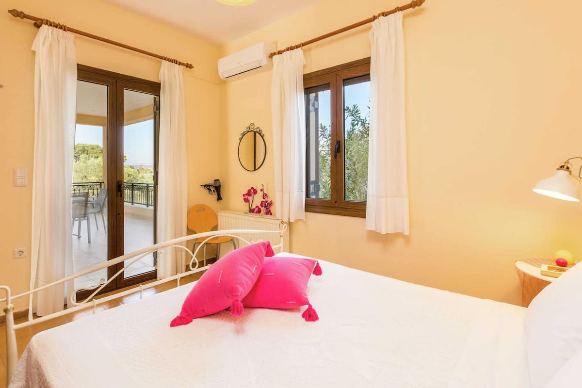 A bedroom with a bed, two pink pillows, a bedside table with a lamp, a window with curtains, an air conditioning unit, and a view of the outdoors.