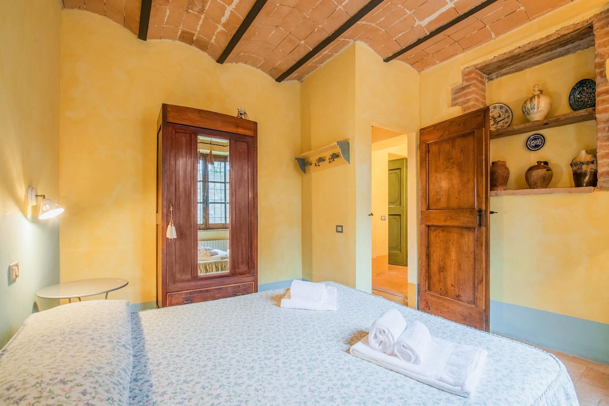 A bedroom with a bed, a wooden door, a shelf with decorative items, and a small table.