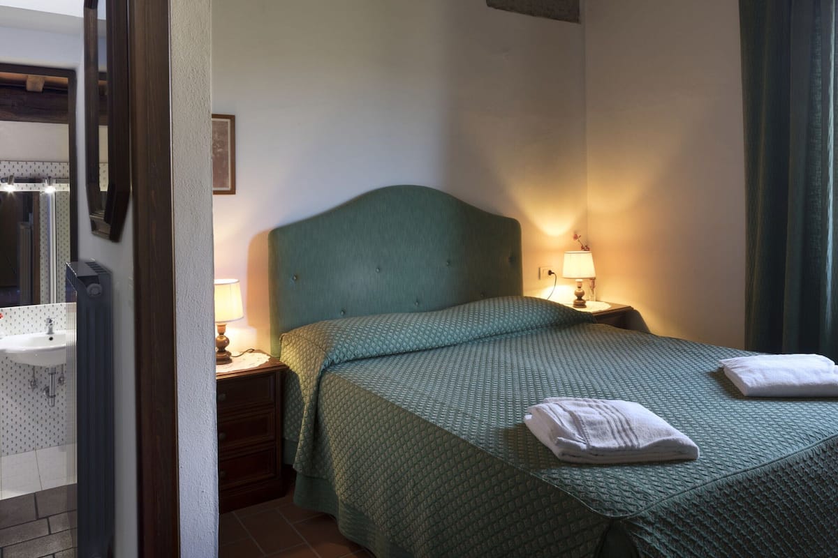 A hotel room with a bed, a bedside table with a lamp, a small nightstand, a mirror, and a bathroom with a sink and shower.