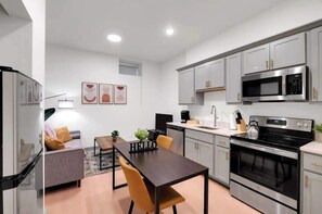 Et kompakt køkken med moderne hårde hvidevarer, et spiseområde med et bord og stole, samt en sofa i et hjørne.