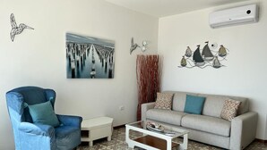 Un salon avec un fauteuil bleu, un canapé beige, une table basse, des carreaux de sol à motifs, un tableau représentant un paysage urbain et des œuvres d’art murales représentant des oiseaux.