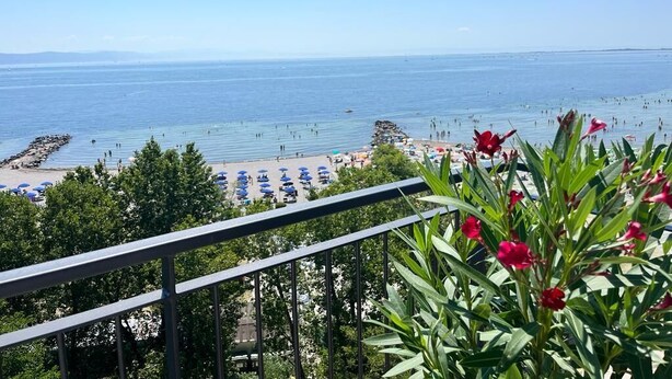 Une vue sur la plage depuis un balcon doté d’une balustrade, donnant sur une plage de sable avec des parasols et des personnes.