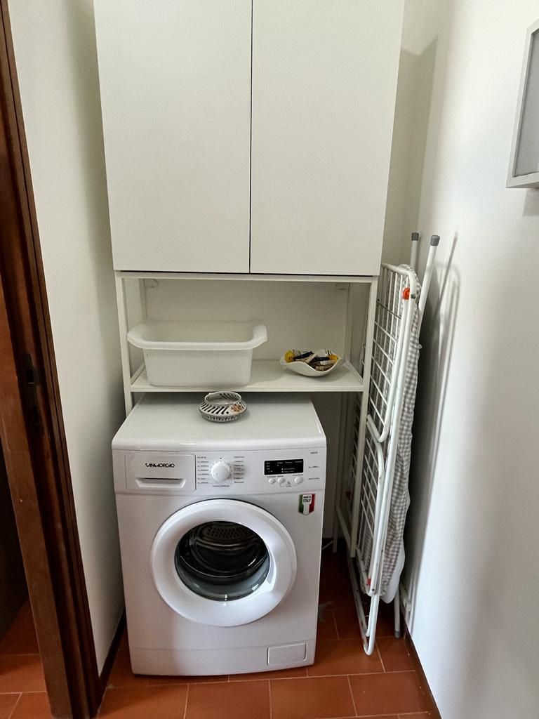 A compact laundry area with a front-loading washing machine, a drying rack, and a small shelf with a basket.