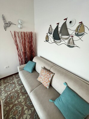 A living room with a beige sofa, patterned cushions, a wall art of sailboats, and a decorative wall piece. - Terrazza sul Mare (Marina Julia)